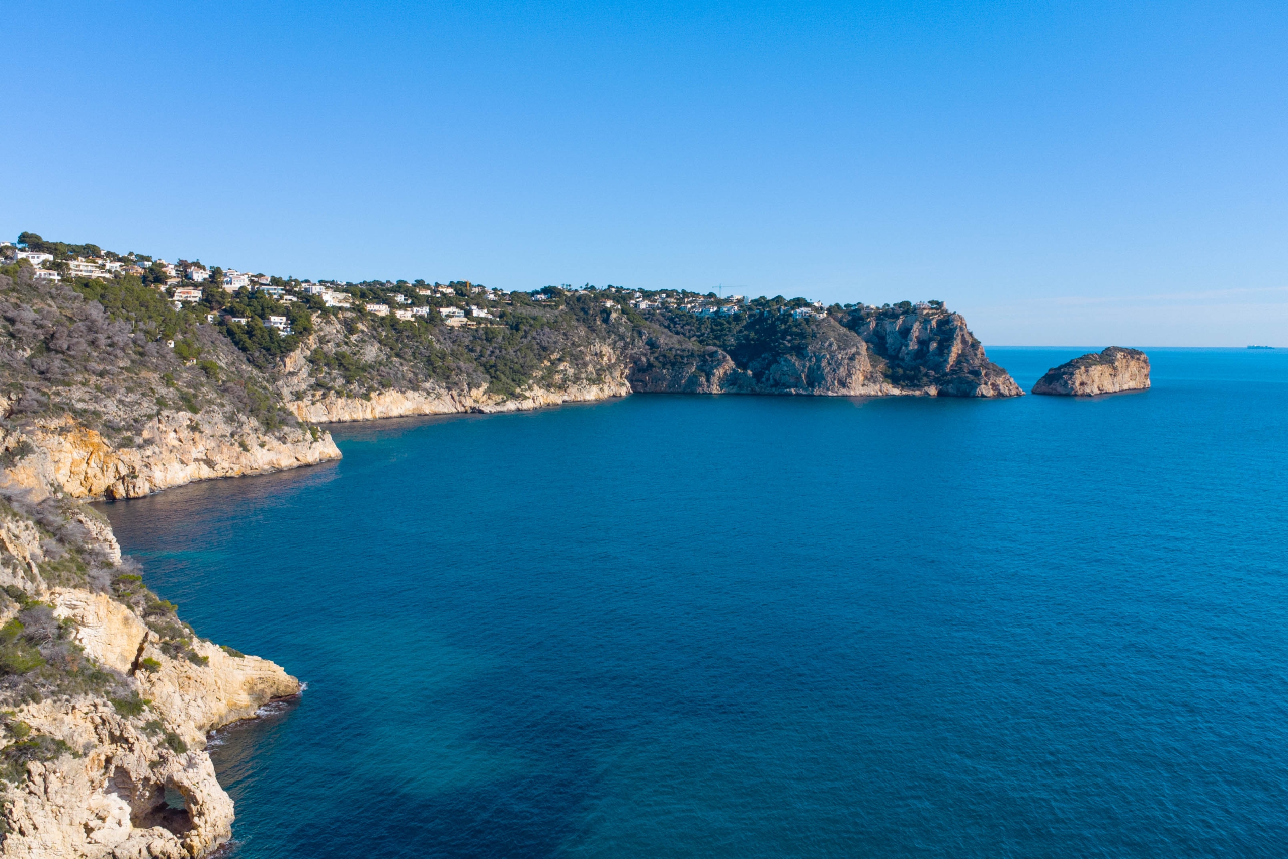 Aerial view of the Jávea coastline featuring limestone cliffs, residential villas overlooking the Mediterranean, and deep blue waters under a clear sky.