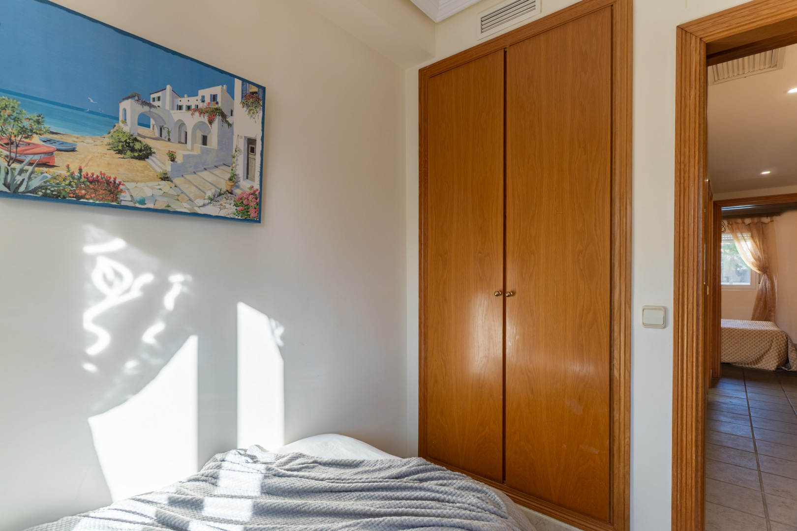 Bedroom featuring a built-in wooden wardrobe and natural sunlight. The doorway with matching trim leads to a tiled hallway and a second bedroom.
