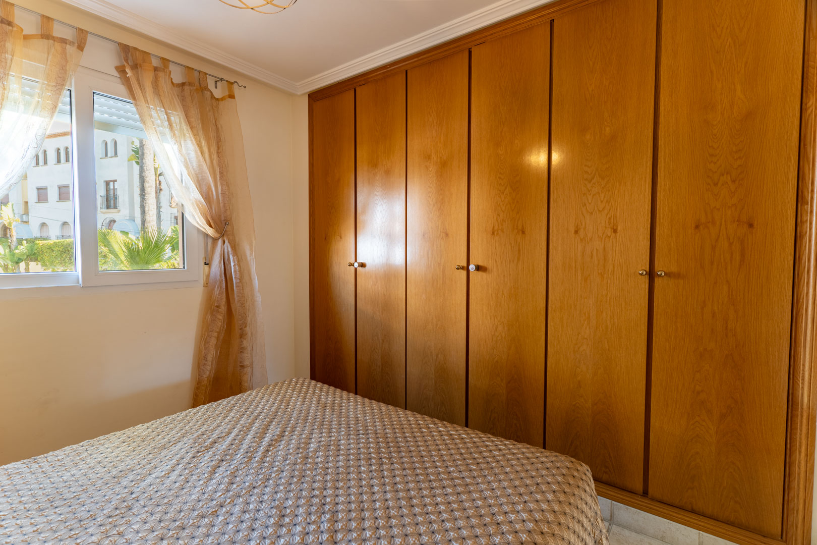 Bedroom featuring a large six-door built-in wooden wardrobe and a window overlooking the landscaped surroundings, offering natural light and storage.