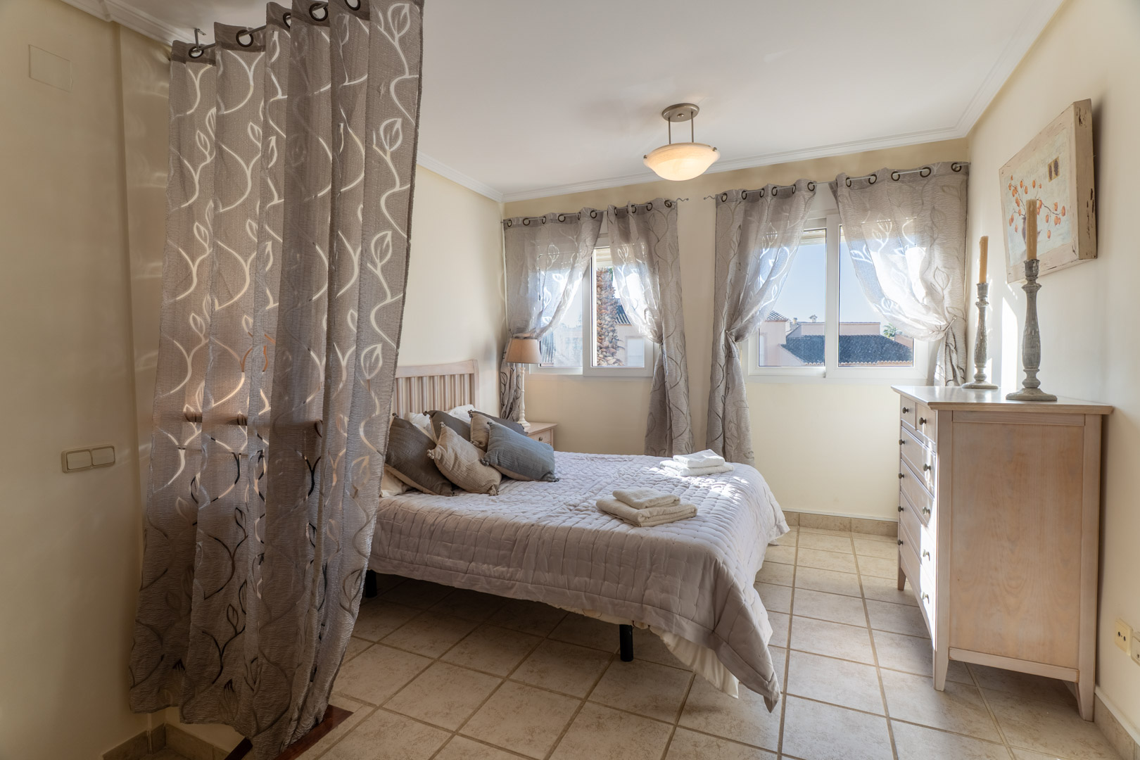 Bedroom featuring tiled flooring, a double bed, wooden dresser, and windows with patterned curtains providing soft natural light.