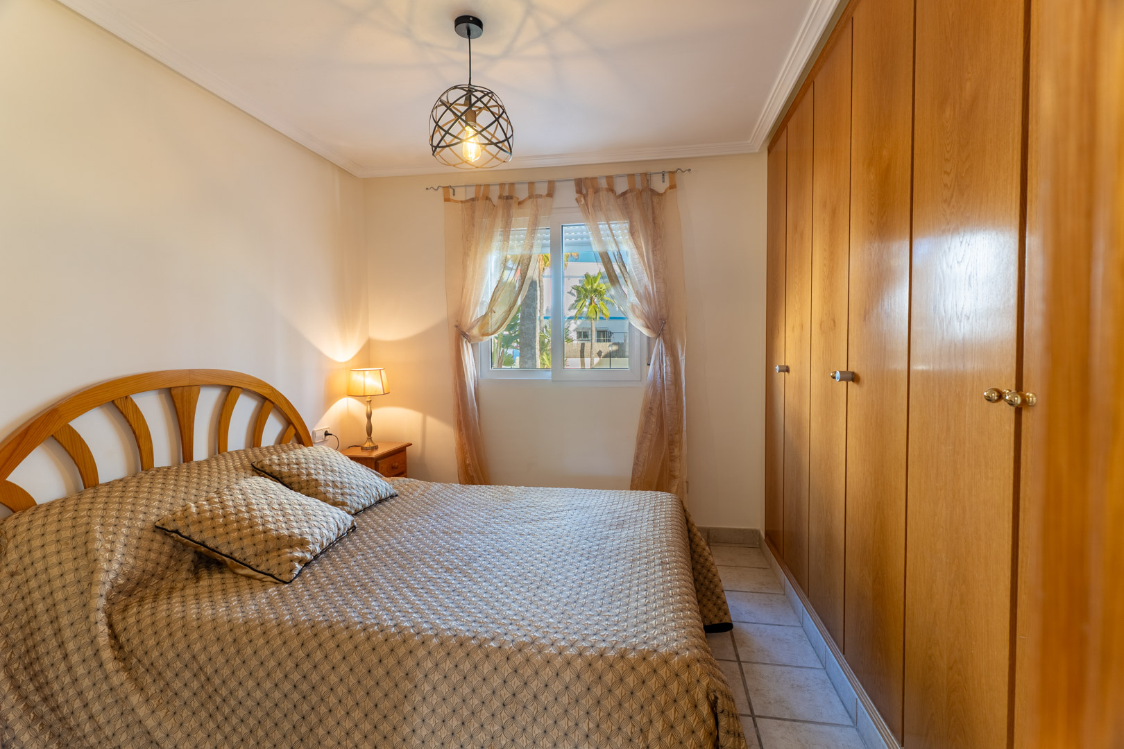 Bright bedroom featuring extensive built-in wooden wardrobes, a double bed, and a window overlooking the community's palm trees and gardens.