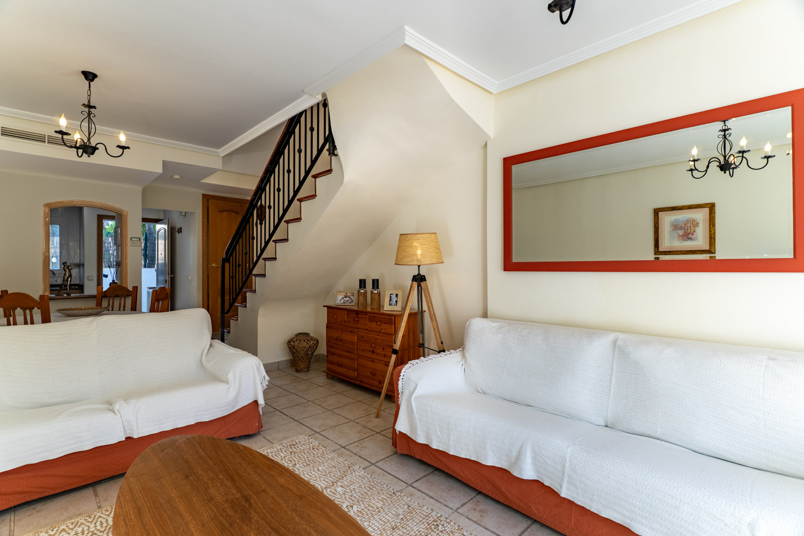 Bright living area featuring tiled floors, a wrought iron staircase, and a large red-framed mirror that enhances the sense of space and light.