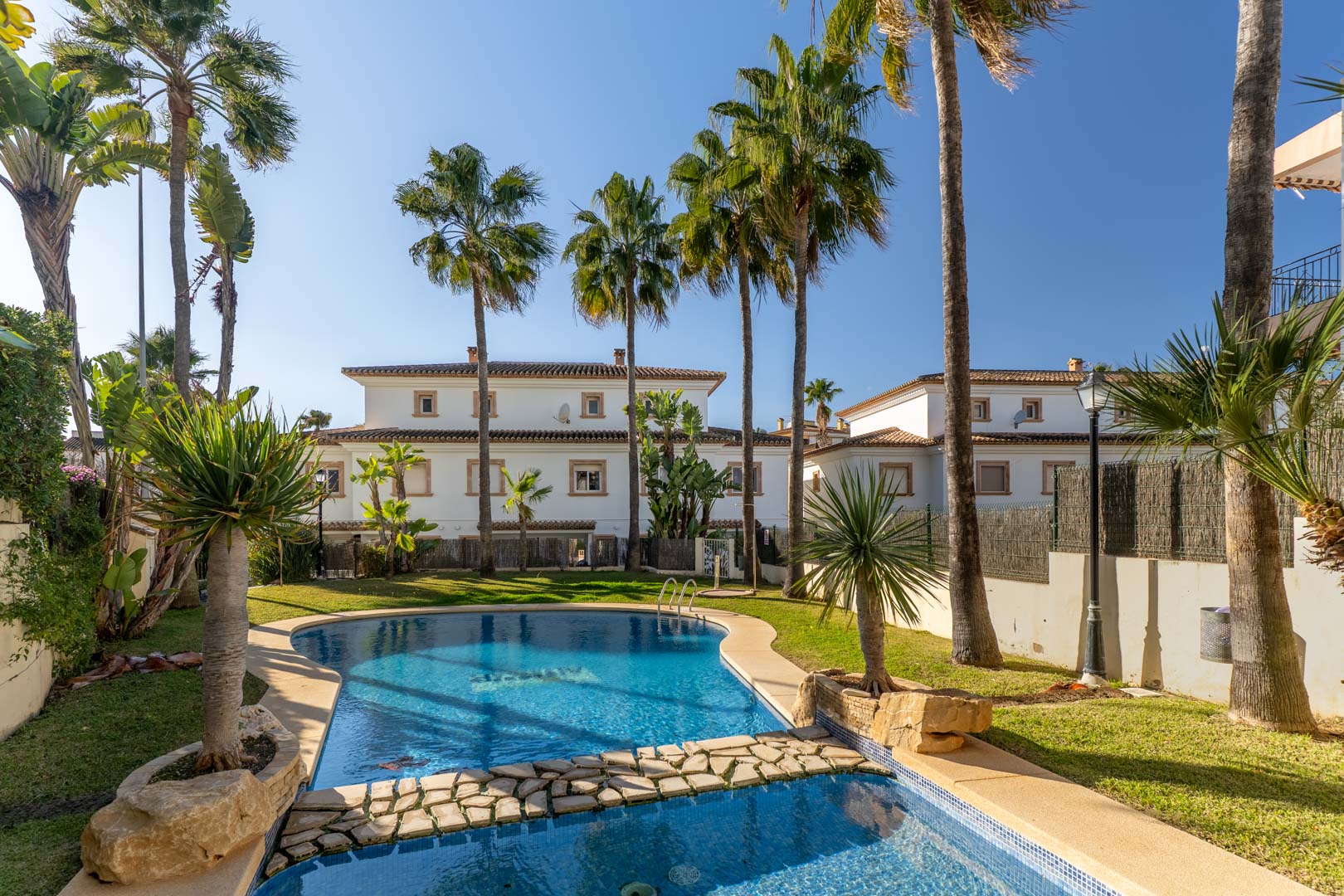 Communal swimming pool featuring blue mosaic tiling and a natural stone bridge, set within landscaped gardens with mature palm trees.