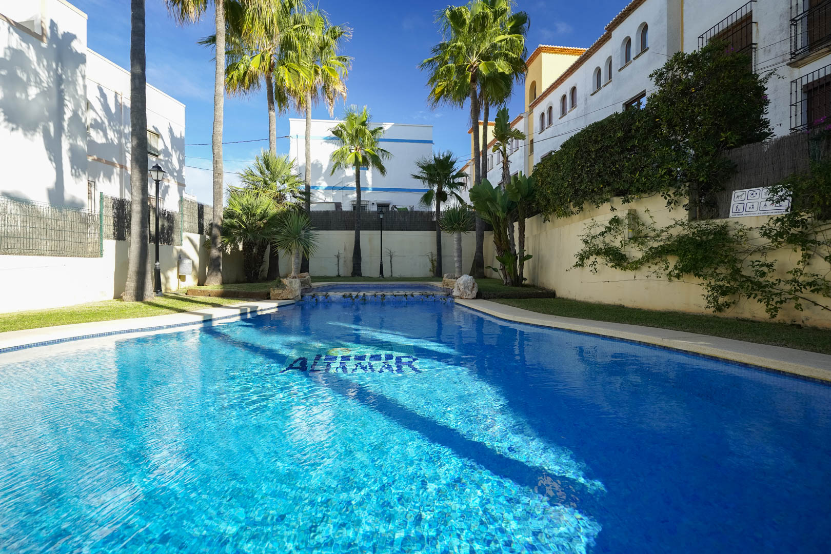 Communal swimming pool with blue tiling, surrounded by mature palm trees and lawn. The area features tropical landscaping and sun-drenched terraces in a secure complex.
