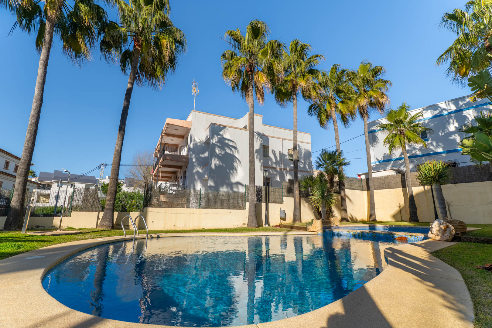 Curved community swimming pool set within manicured lawns and flanked by mature palm trees, offering a private outdoor space under the Mediterranean sun.