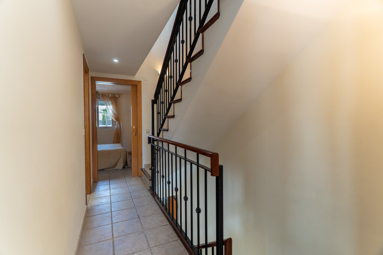 Distribution hallway with ceramic tiling leading to bedrooms, featuring a staircase with a wooden handrail and black wrought-iron balusters.