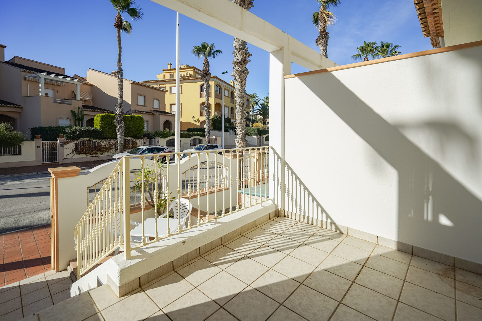 Elevated front terrace featuring ceramic tiling and metal railings, offering views of the palm-lined street and surrounding Mediterranean architecture.