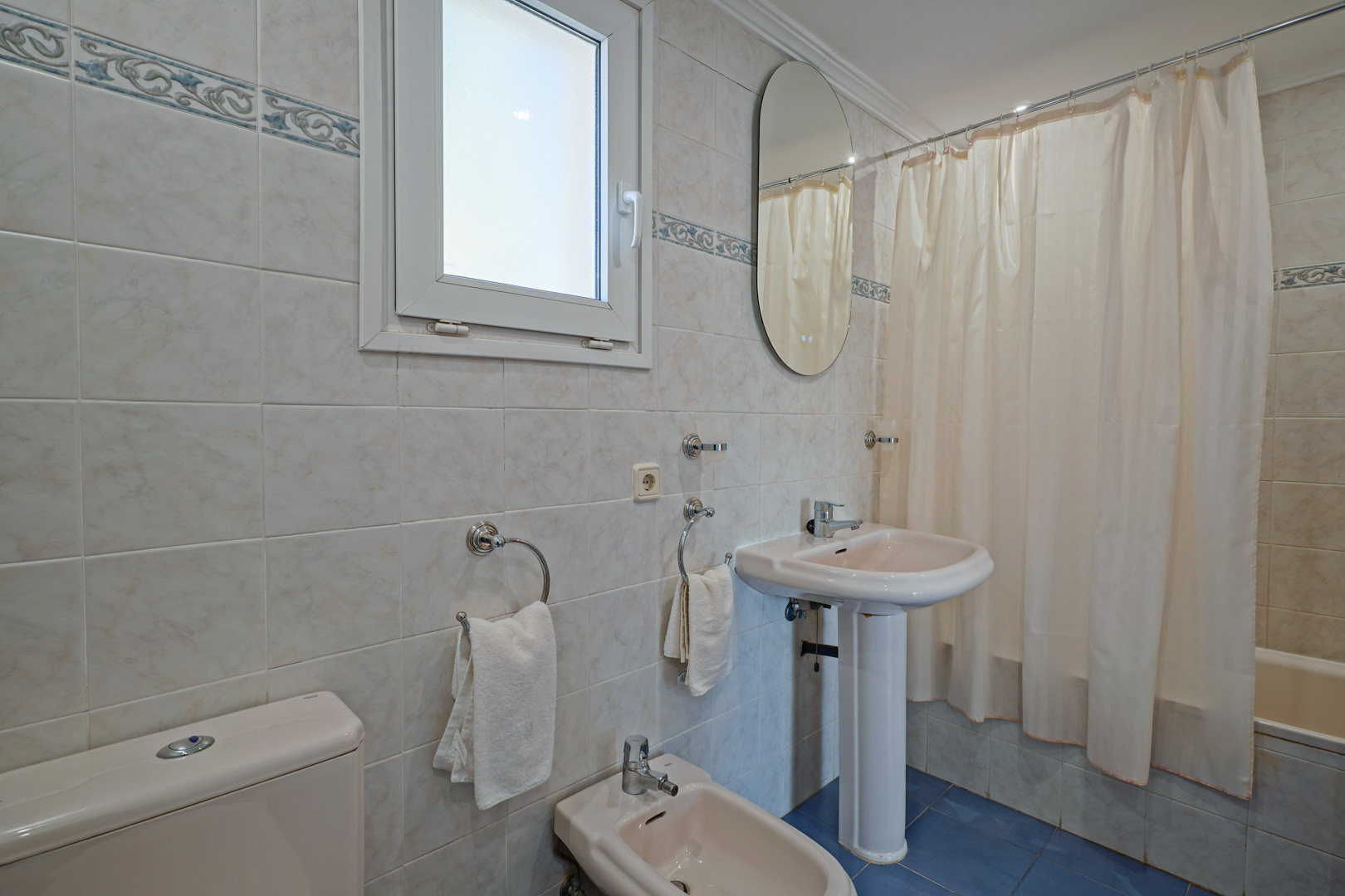 Family bathroom featuring cream-colored fixtures, a pedestal sink, and a bidet. It includes a bathtub with a curtain, blue floor tiles, and a frosted window for natural light.