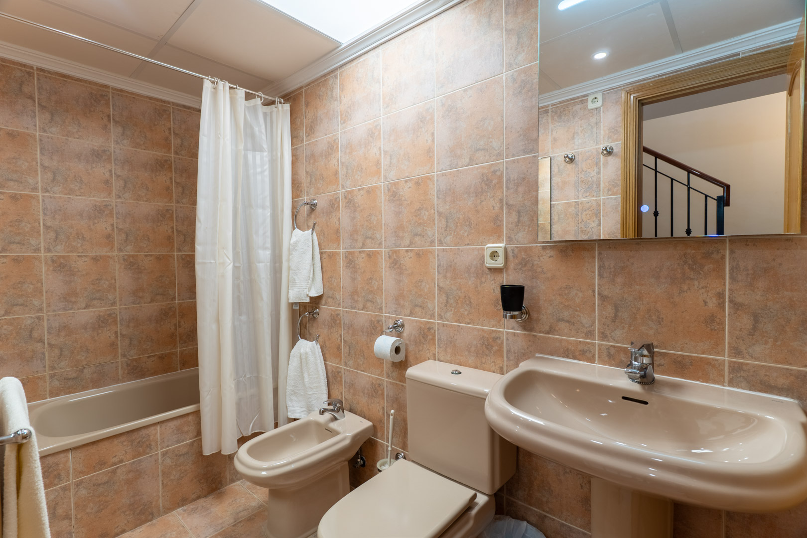 Full bathroom featuring a bathtub with curtain, bidet, pedestal sink, and earth-toned ceramic tiling illuminated by a skylight.