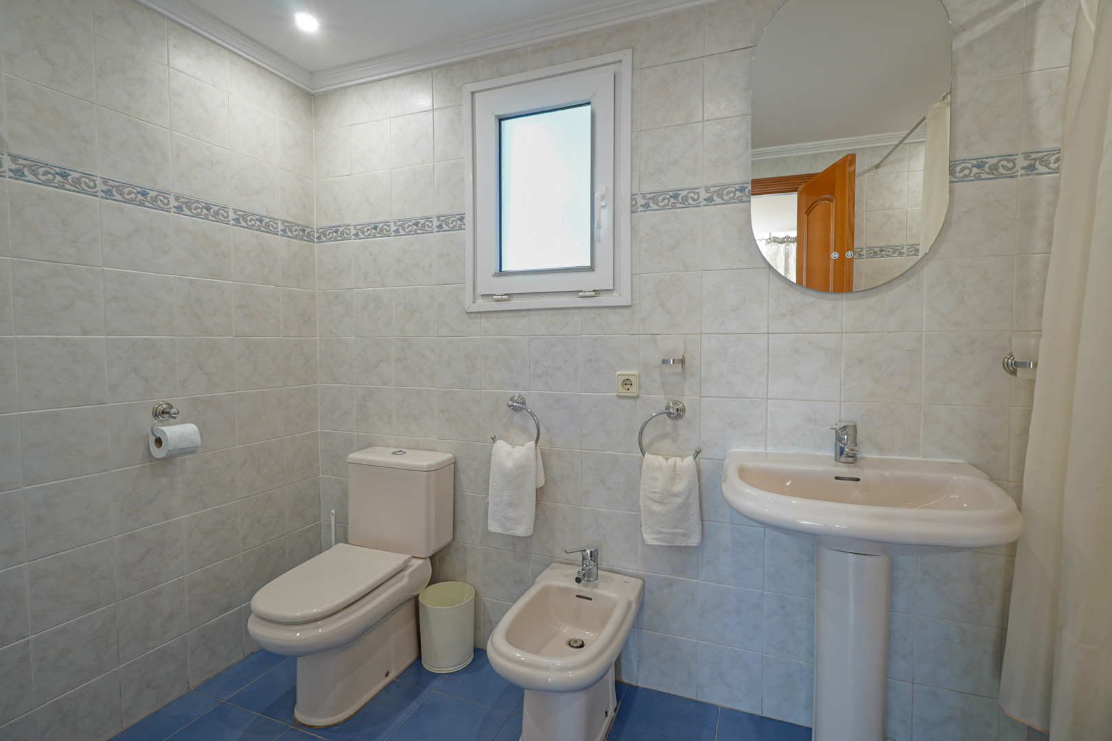 Full bathroom featuring light-toned wall tiles with a decorative border and blue flooring. Equipped with a toilet, bidet, pedestal sink, and a frosted window.