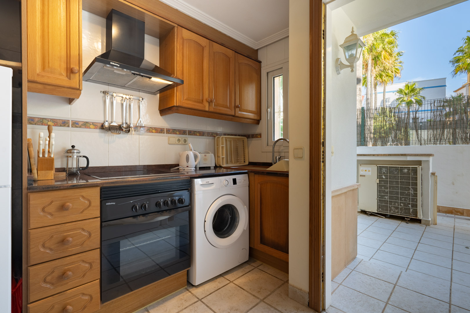 Functional kitchen featuring wooden cabinetry, integrated appliances, and a washing machine, with direct access to a private tiled patio and outdoor views.