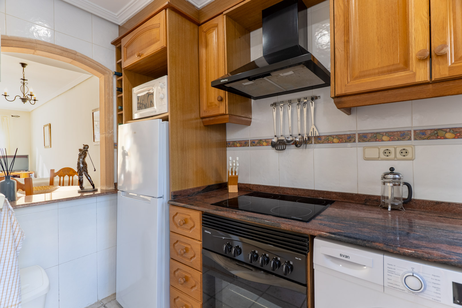 Functional kitchen with wooden cabinets, granite countertops, and appliances, featuring a stone-framed archway overlooking the living area.
