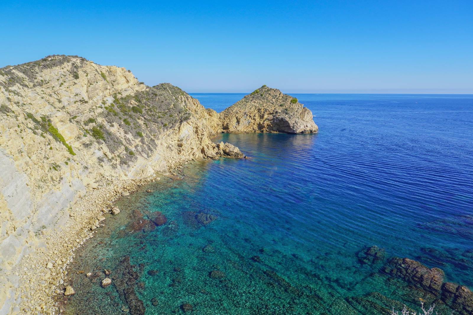 Panoramic view of the Jávea coastline featuring limestone cliffs and crystal-clear turquoise Mediterranean waters under a clear blue sky.