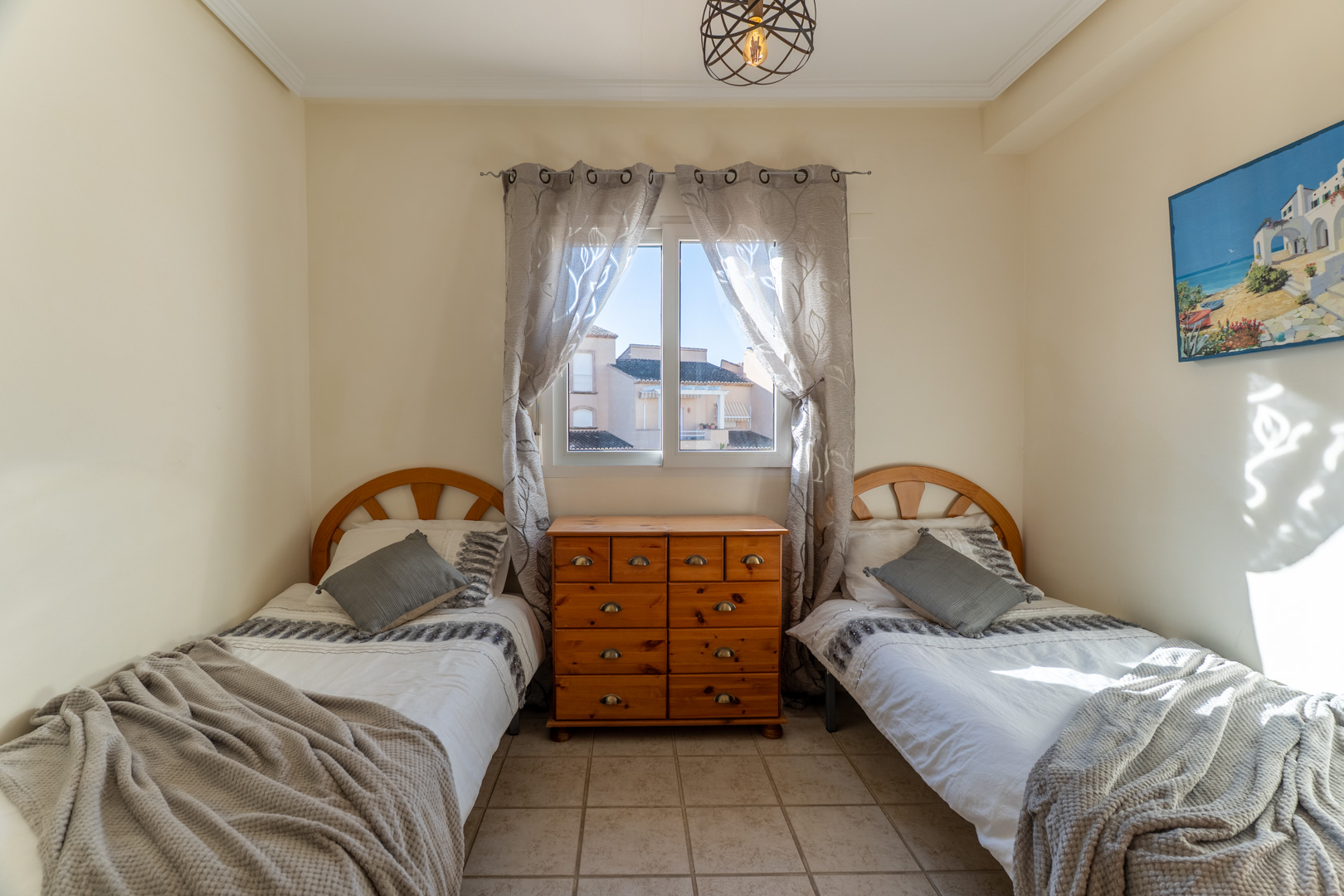 Twin bedroom featuring wooden headboards and a central pine chest of drawers. The space includes tiled flooring, natural light, and a modern wire-frame pendant.