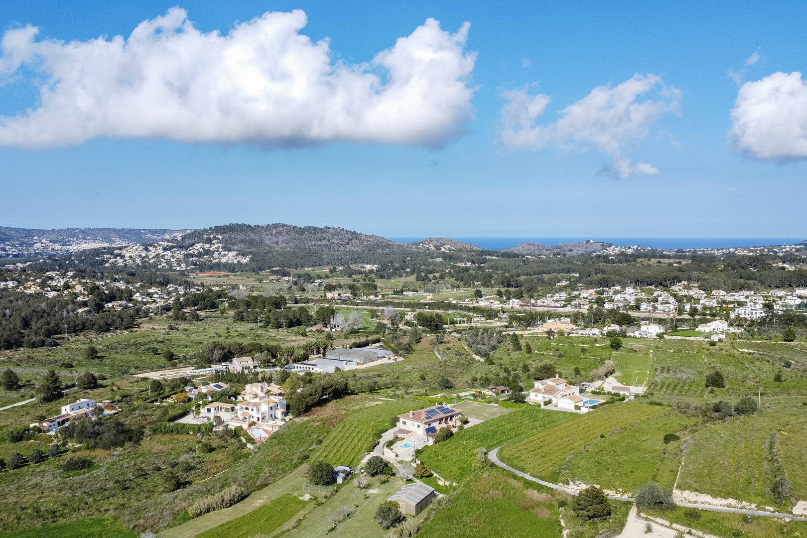 Aerial panoramic view of the Mediterranean landscape featuring green valleys, scattered villas with swimming pools, and distant sea views under a bright sky.