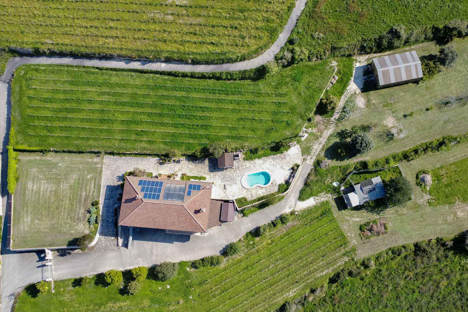 Aerial view of a rural property featuring a main residence with solar panels, a kidney-shaped swimming pool, and organized rows of crops on a large green plot.