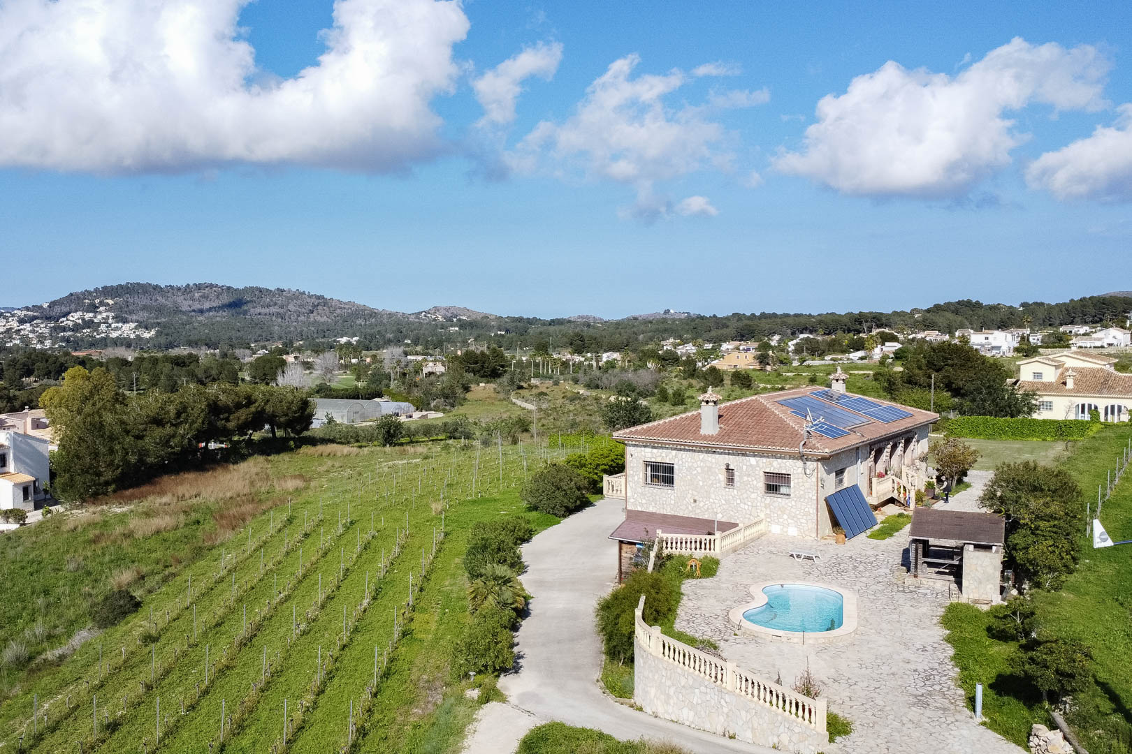 Aerial view of a stone country house featuring solar panels, a kidney-shaped pool with stone terrace, and adjacent vineyard rows set against a hilly landscape.