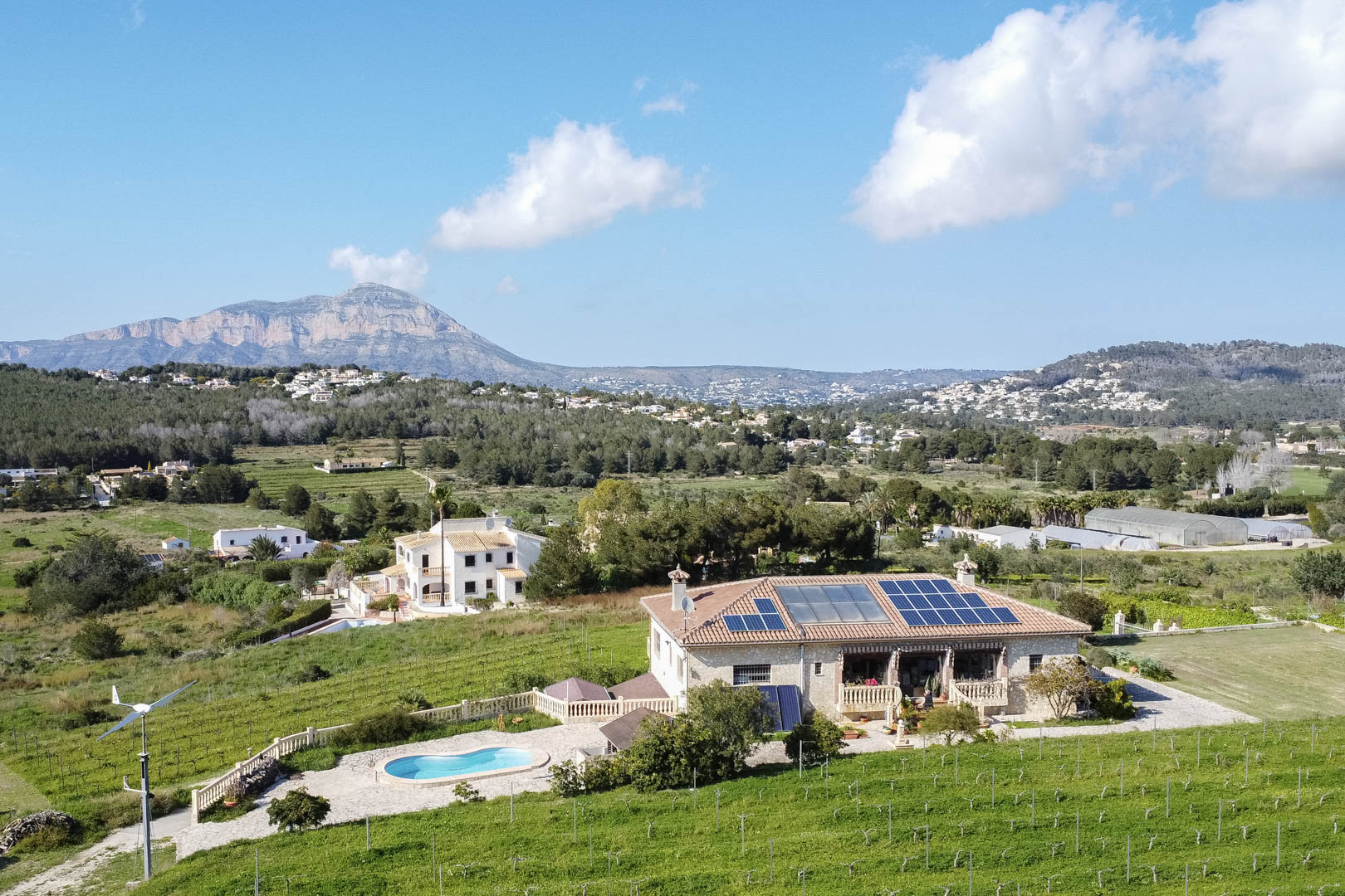 Aerial view of a stone country house with solar panels, a wind turbine, and a kidney-shaped pool, surrounded by vineyards and a mountain landscape.