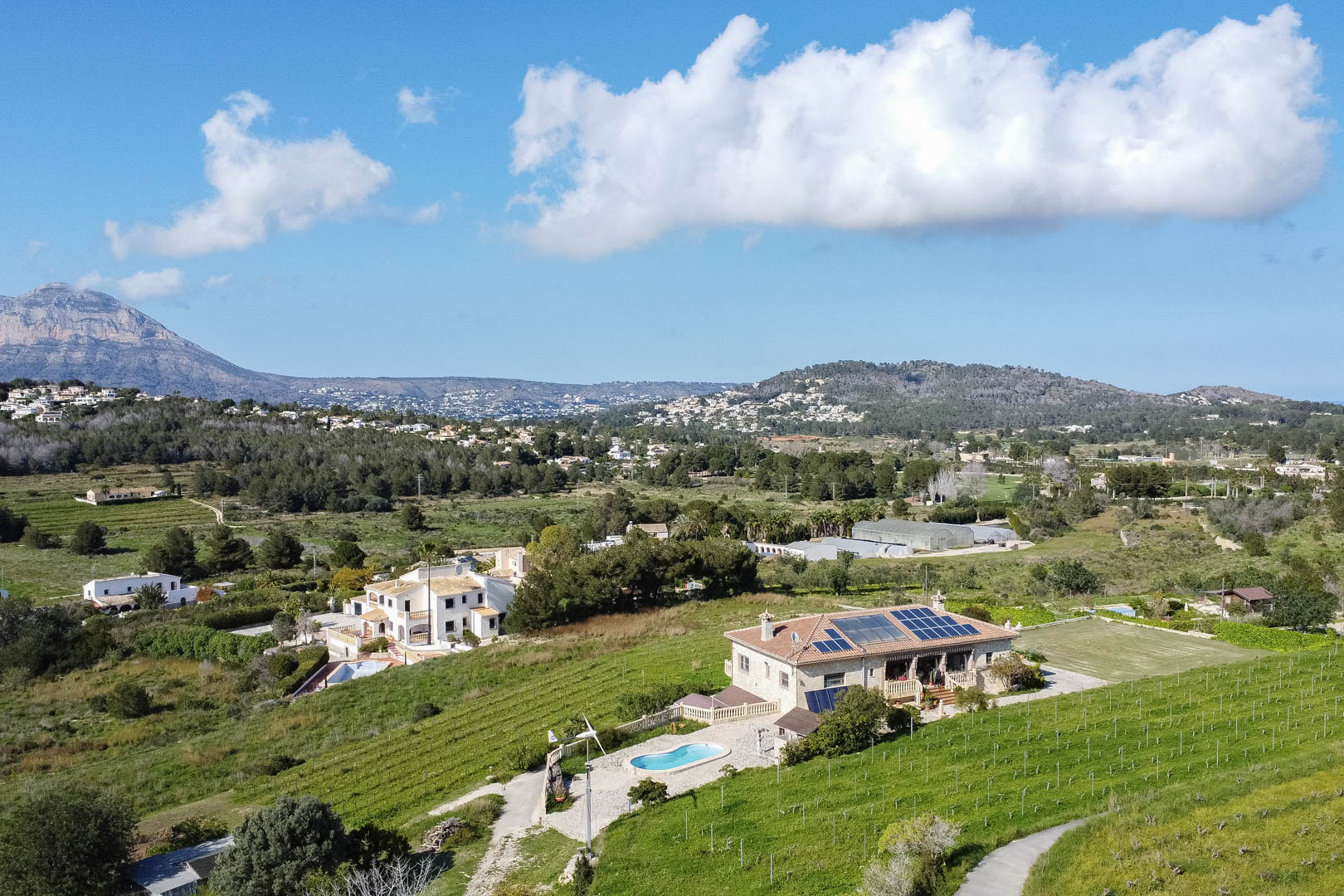 Aerial view of a stone villa featuring rooftop solar panels, a private pool, and a wind turbine, surrounded by lush vineyards and a mountainous backdrop.