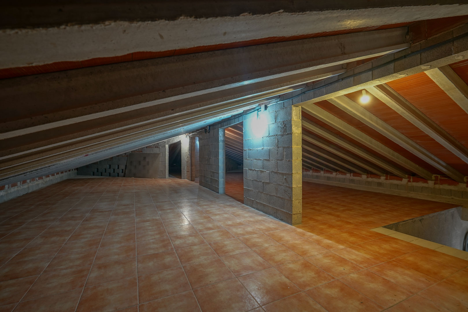 Attic storage area featuring terracotta-colored floor tiles, exposed concrete ceiling beams, and structural cinder block walls with integrated lighting.