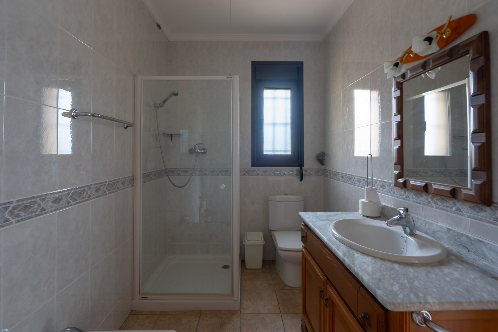 Bathroom featuring a marble-topped vanity, wooden cabinetry, and a walk-in shower with glass enclosure. Tiled walls include a decorative border and a frosted window.