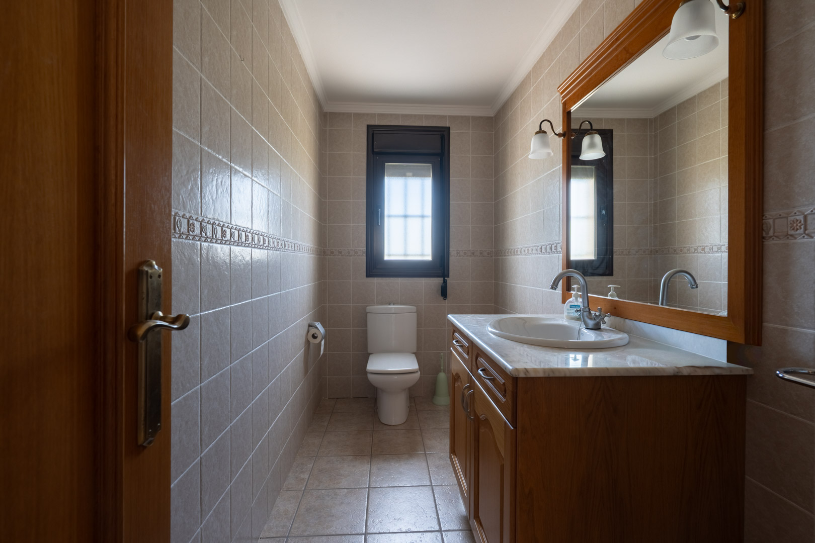 Bathroom featuring a wooden vanity with a marble countertop and a large framed mirror. Beige tiled walls and a small window create a functional space.