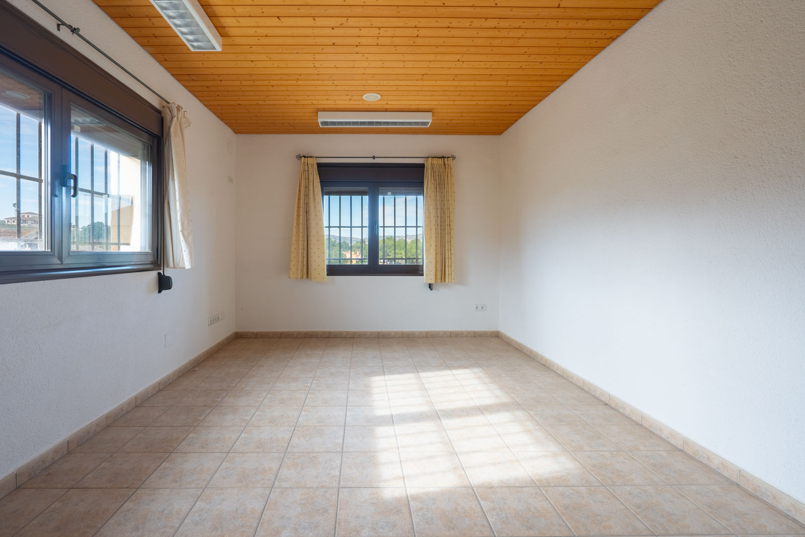 Bright empty room with a natural wood paneled ceiling, beige tiled flooring, and two windows featuring security bars and cream curtains.