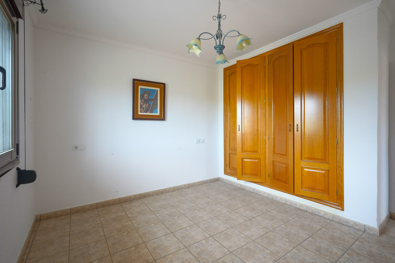 Empty room featuring a large built-in wooden wardrobe with four doors, beige ceramic tile flooring, and a decorative floral chandelier against white walls.
