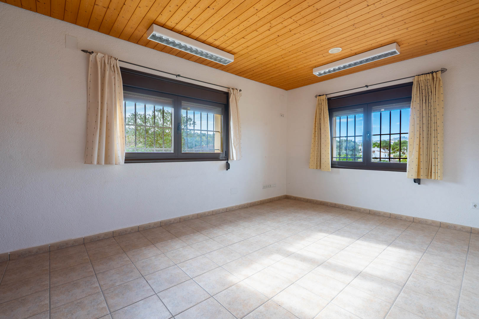 Empty room featuring a natural wood-paneled ceiling, ceramic tile flooring, and dual windows with security bars and beige curtains.