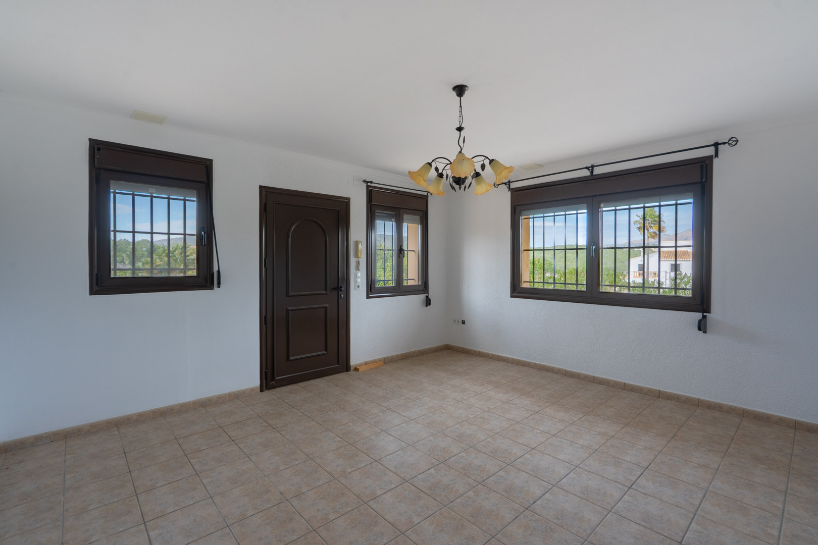 Empty room featuring beige tiled flooring, dark brown window frames with security bars, and a floral chandelier. Large windows offer views of the countryside.
