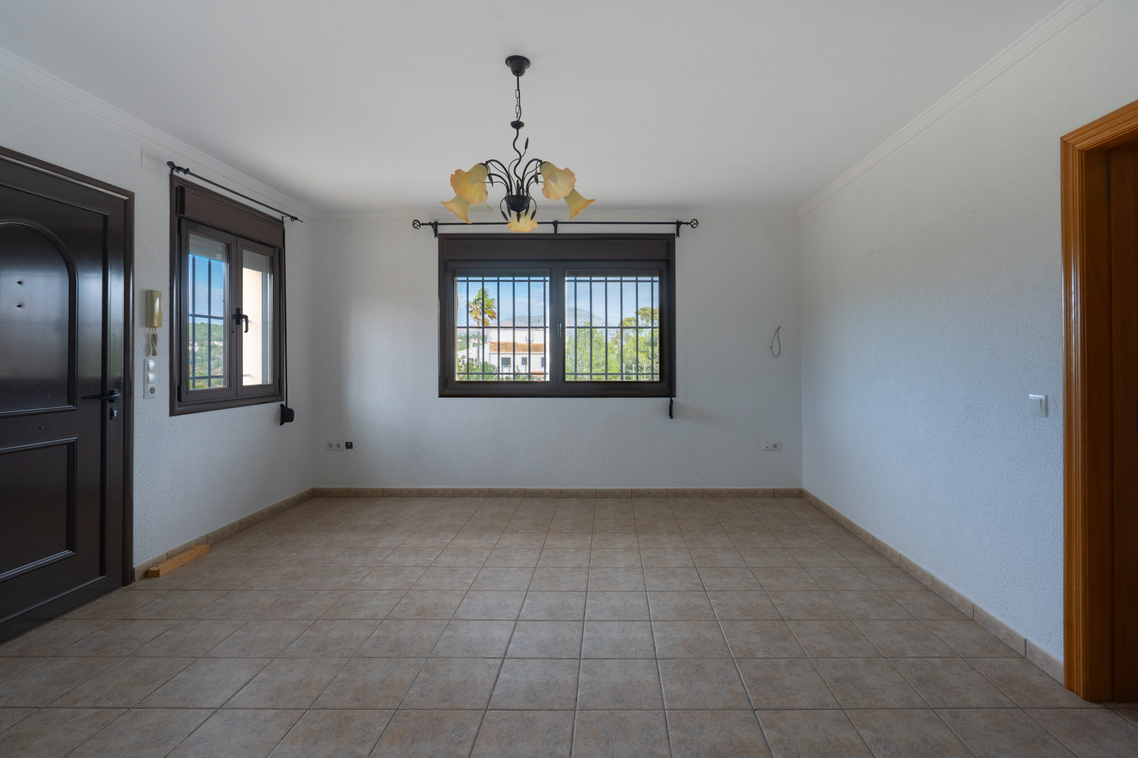Empty room with beige ceramic floor tiles, dark-framed windows with security bars, and a wrought iron chandelier with floral glass shades.