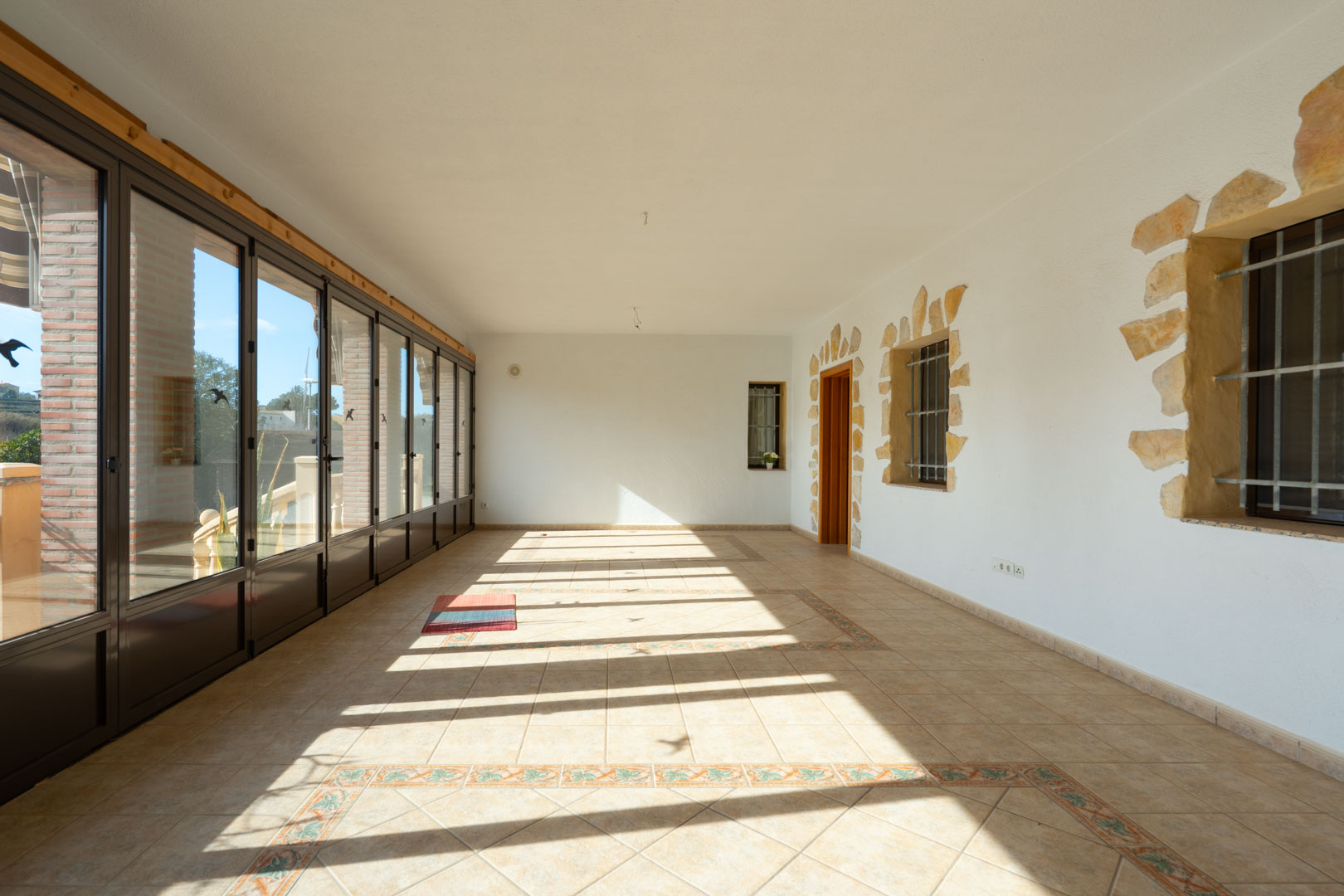 Enclosed sunroom featuring large glass panels, decorative tiled flooring, and white walls accented with natural stone around the window frames.