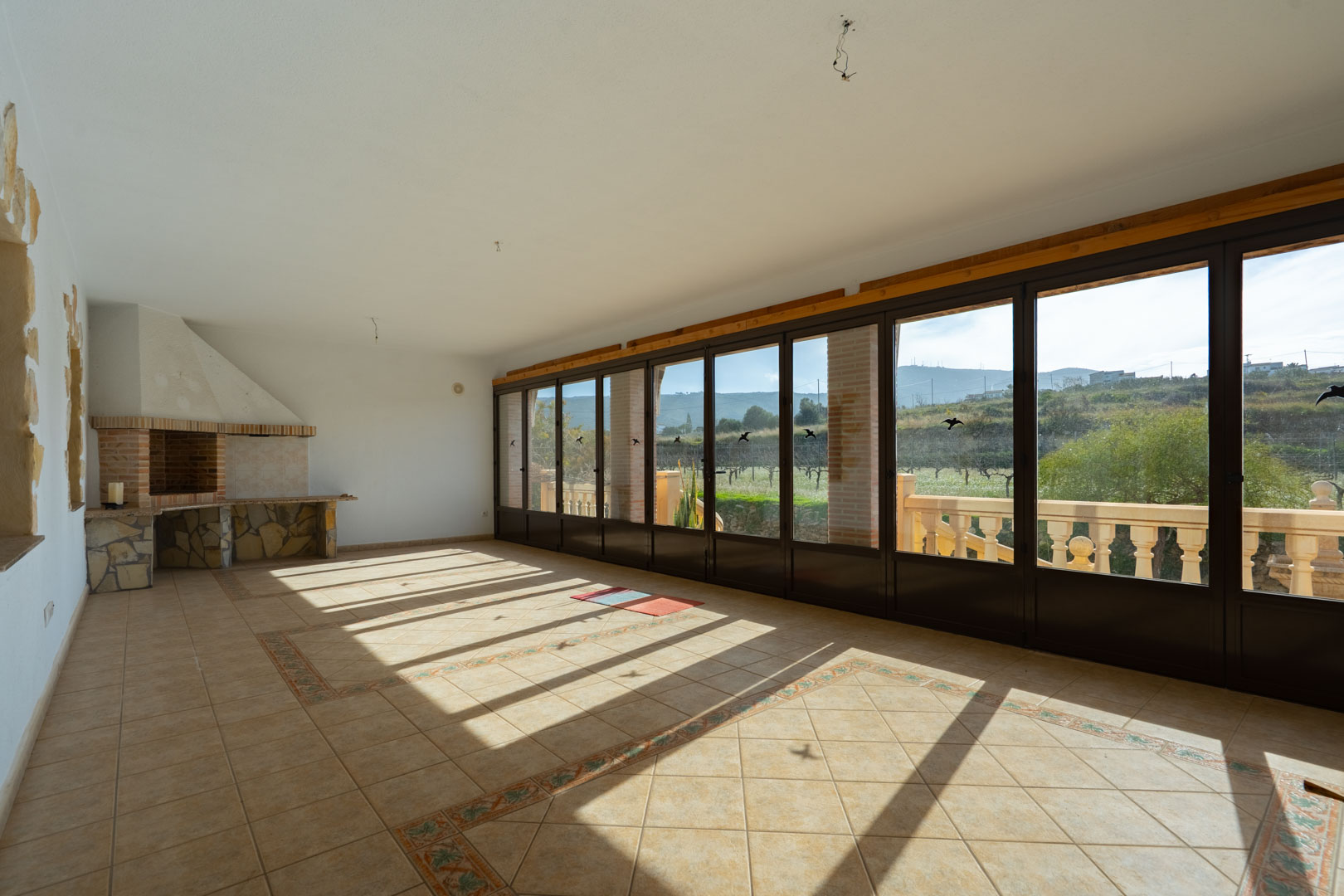 Enclosed terrace with a stone-clad barbecue, tiled floors with decorative borders, and floor-to-ceiling glass doors overlooking the hills.