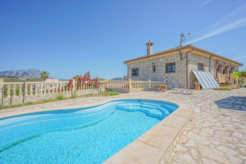 Exterior view featuring a curved swimming pool and a stone-clad villa. The terrace is paved with natural stone, offering views of the distant mountains.
