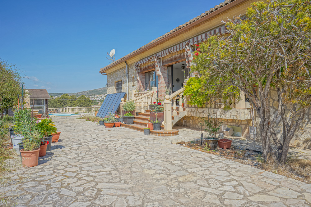 Exterior view featuring a stone-paved patio, natural stone facade, and solar panels. Stairs with balustrades lead to a terrace with striped awnings.