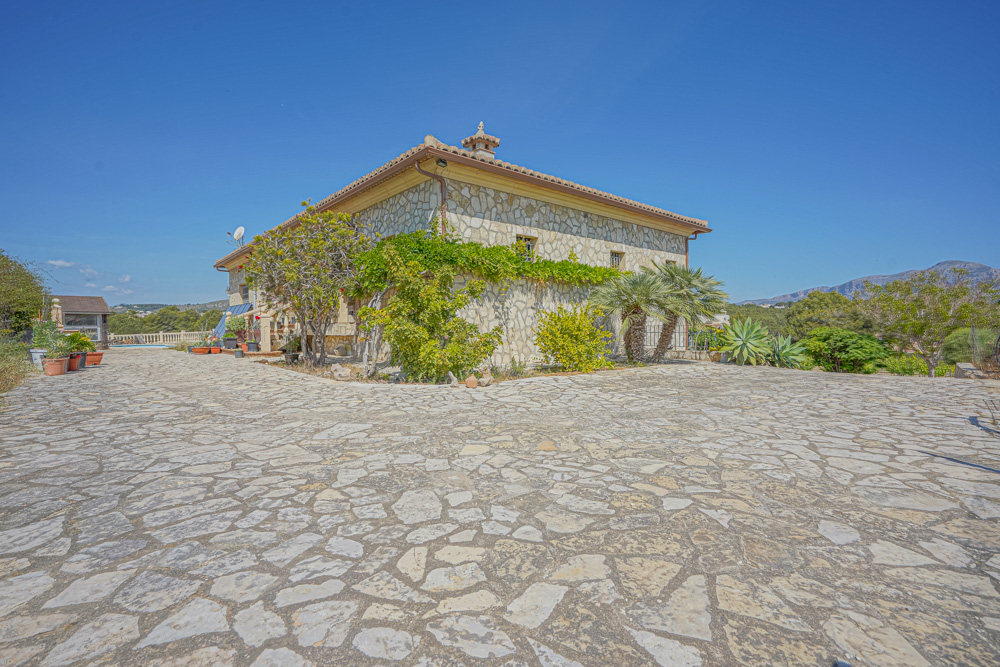Exterior view of a stone-clad villa featuring a large natural stone-paved driveway, Mediterranean landscaping with palm trees, and a terracotta tiled roof.