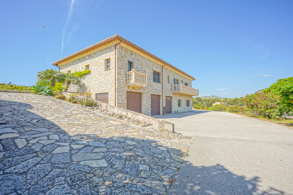 Exterior view of a stone-clad villa featuring a triple garage, stone-paved driveway, and Mediterranean landscaping under a clear blue sky.