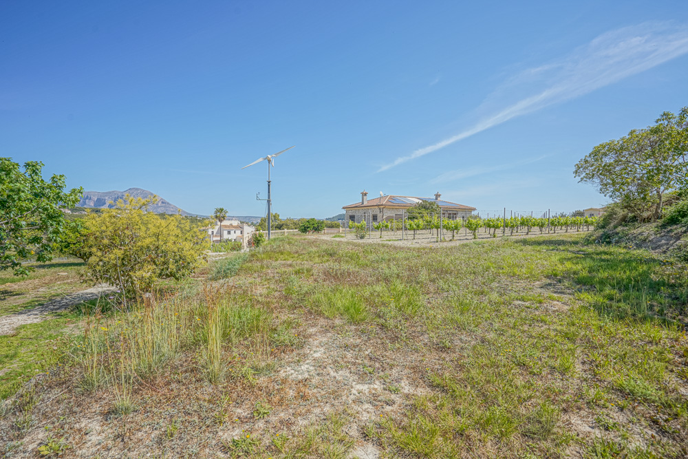 Exterior view of a stone villa with solar panels and a wind turbine, featuring a private vineyard and distant mountain views under a clear sky.