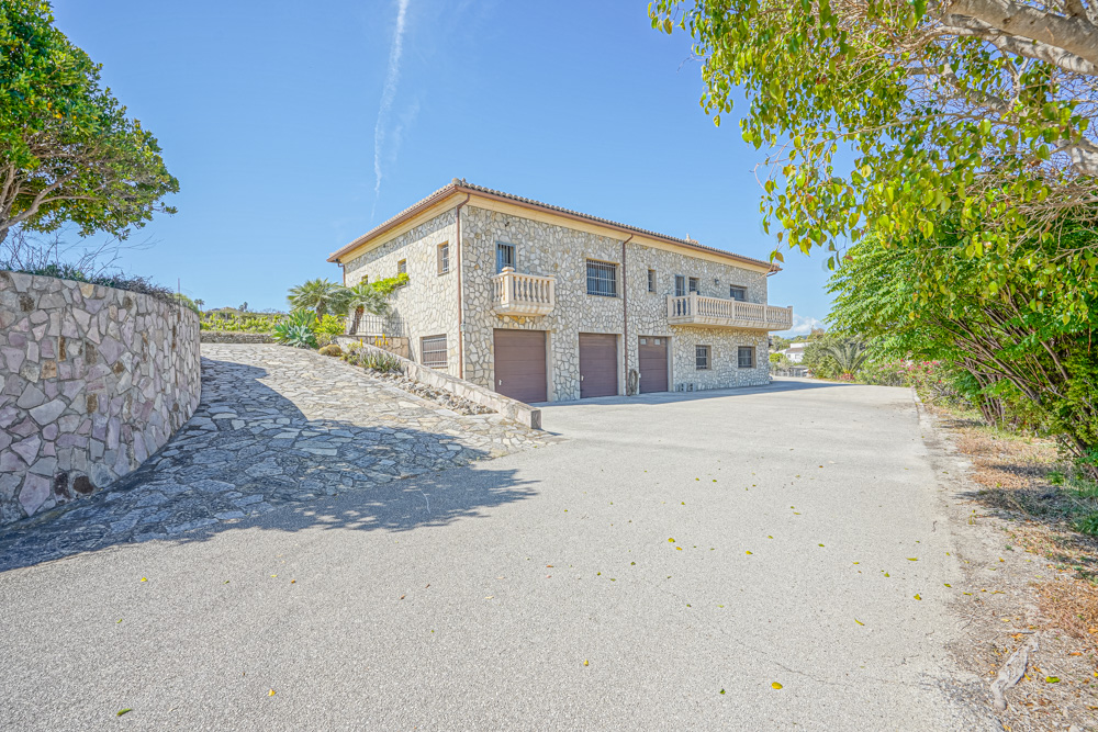 Exterior view of a two-story stone villa featuring three garage bays, a stone-paved driveway, and balconies with classic balustrades.