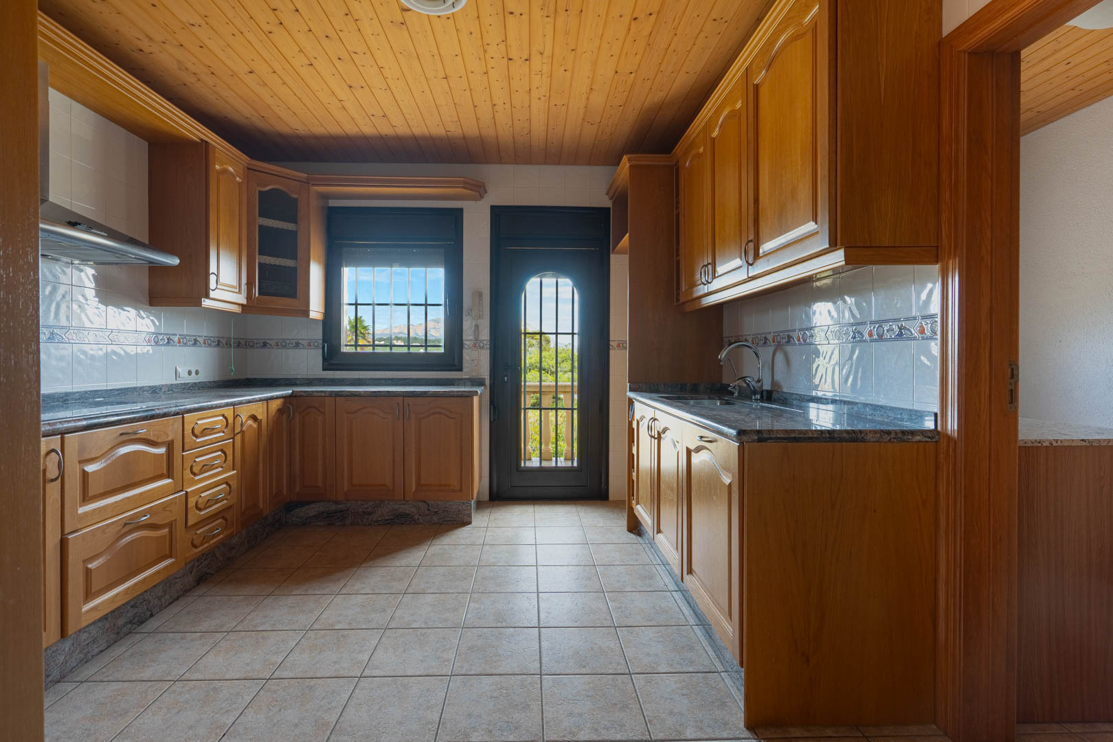 Kitchen with wooden cabinetry and granite countertops, featuring a wood-paneled ceiling, tiled flooring, and a door leading to an outdoor balcony with mountain views.