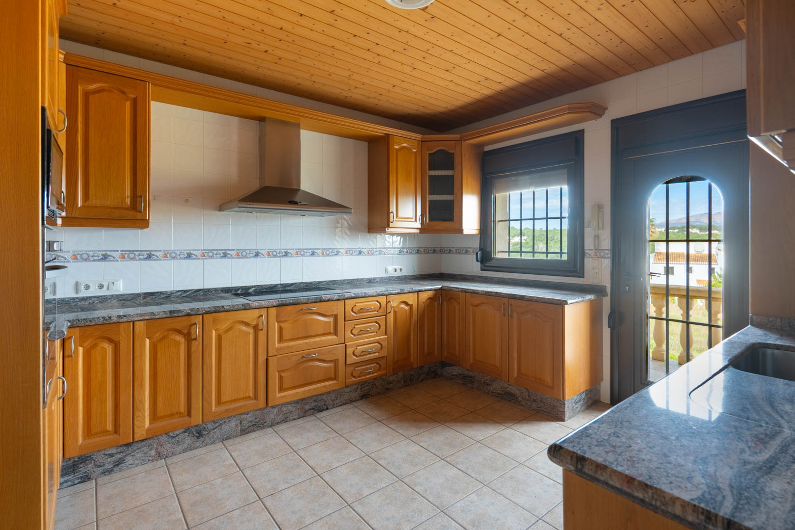 Kitchen with wooden cabinetry, granite countertops, and a wood-paneled ceiling. Features a stainless steel extractor and access to a terrace with landscape views.