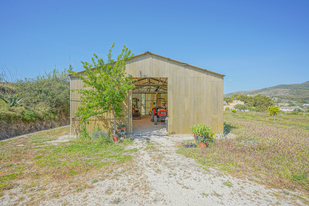 Large corrugated metal agricultural warehouse with a gabled roof, housing a tractor and machinery, situated on a rural plot with mountain views.