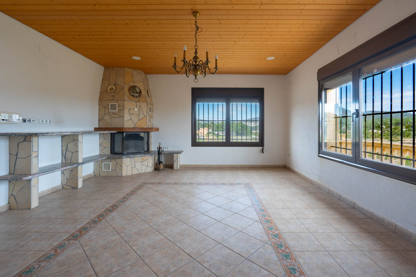 Living room featuring a wood-paneled ceiling, stone-clad fireplace, and tiled flooring with a decorative border. Large windows offer rural views.