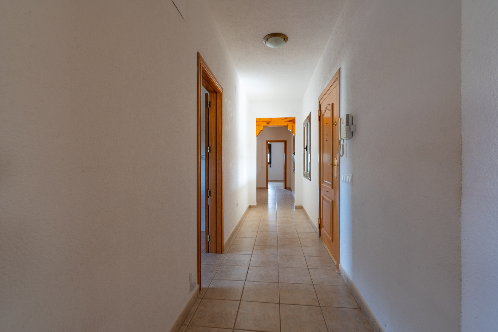 Long hallway featuring neutral ceramic tile flooring, white walls, and light wood doors. An intercom is mounted near the entrance, leading to a decorative wooden arch.