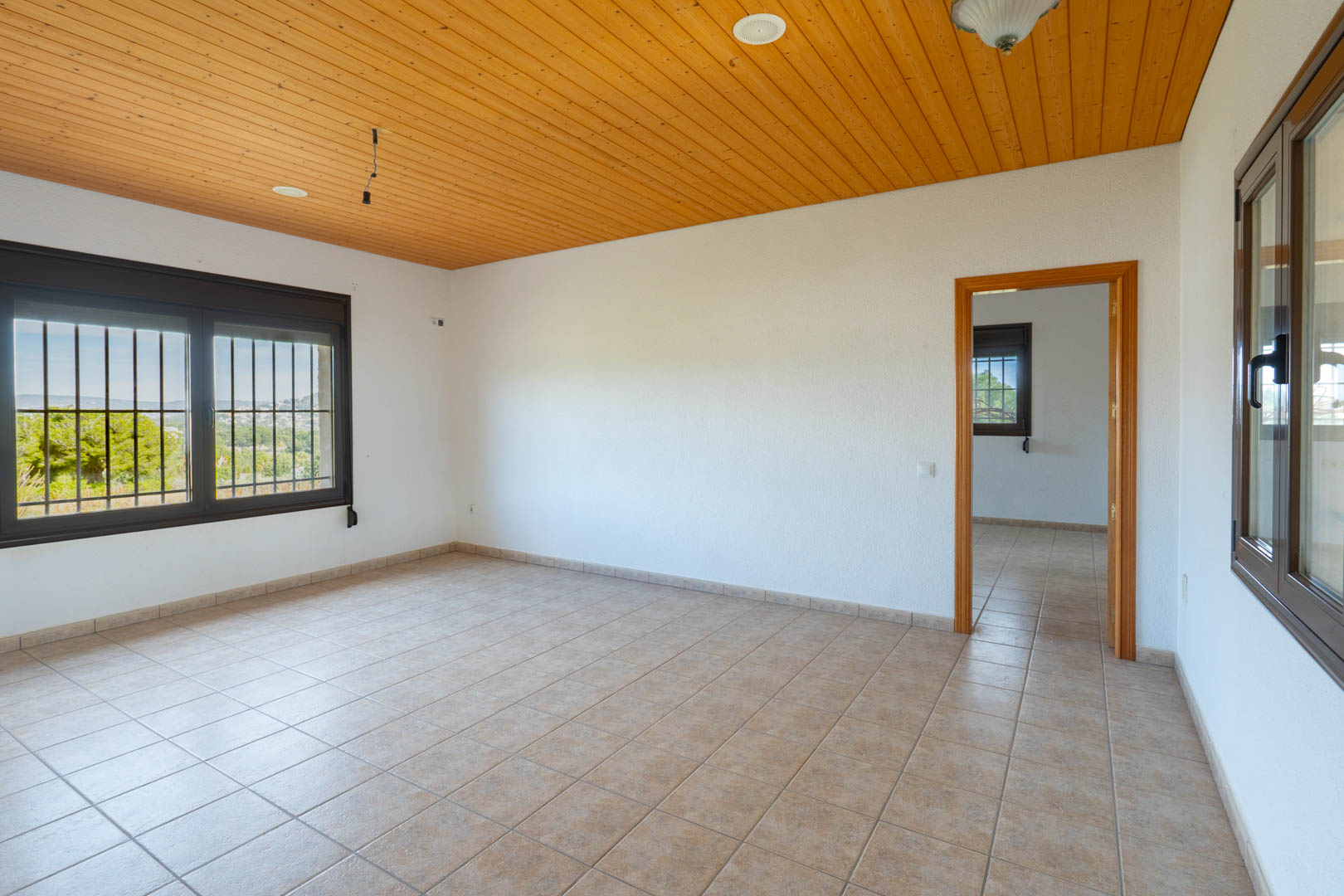 Room featuring a wooden paneled ceiling, beige tile flooring, and dark-framed windows with security bars overlooking the surrounding landscape.