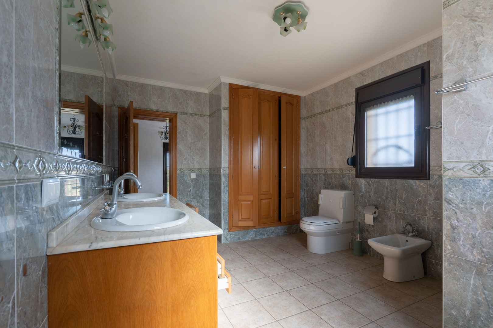Spacious bathroom featuring a double marble vanity, integrated wooden wardrobes, and full wall tiling with decorative borders. Includes a toilet and bidet.