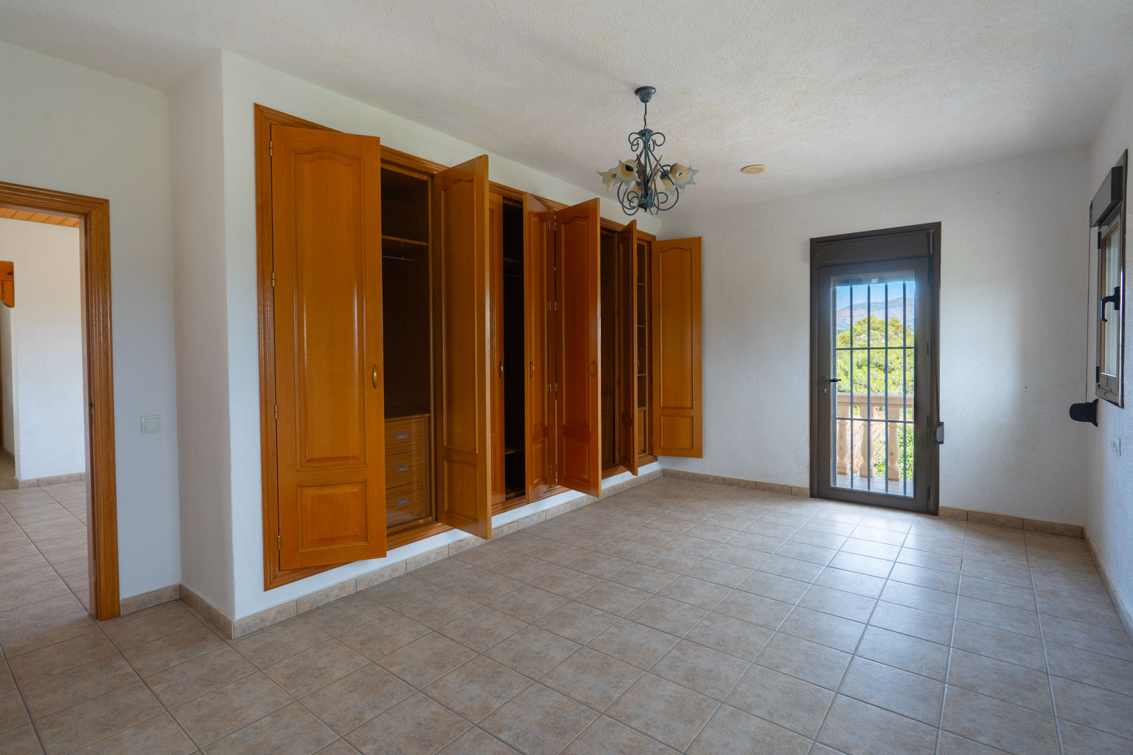 Spacious bedroom featuring a large built-in wooden wardrobe, tiled flooring, and a glass door providing access to a balcony with views of the landscape.