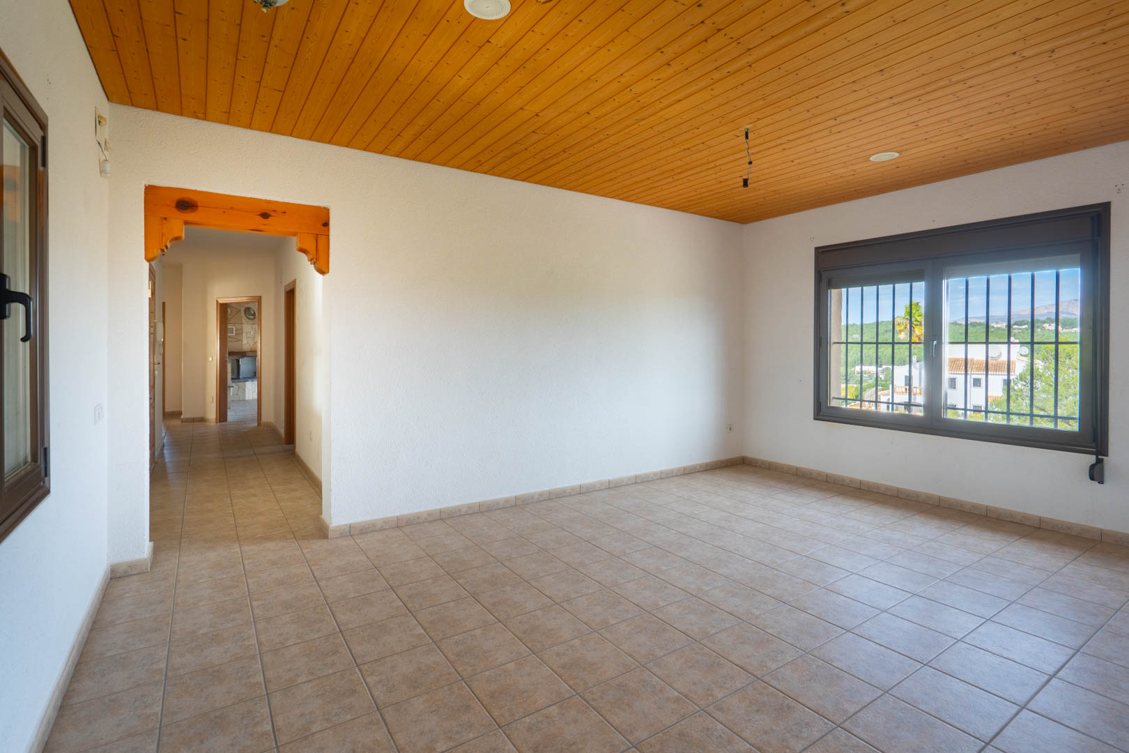 Spacious empty room featuring a pine wood paneled ceiling, tiled floors, and a window with security bars overlooking the surrounding landscape.