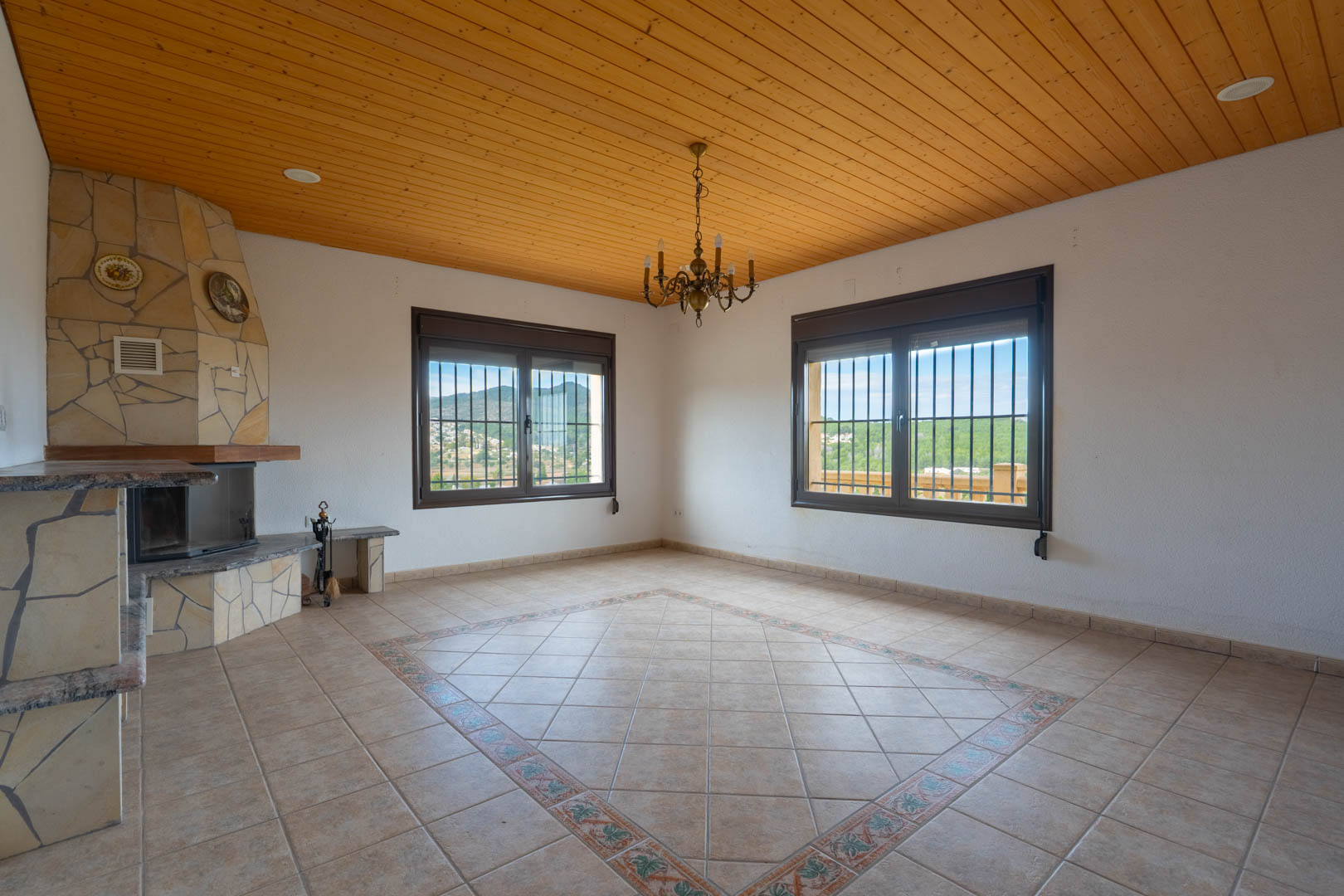 Spacious living area featuring a wood-paneled ceiling, a corner stone fireplace, and tiled flooring with a decorative central mosaic. Large windows offer views of the hills.