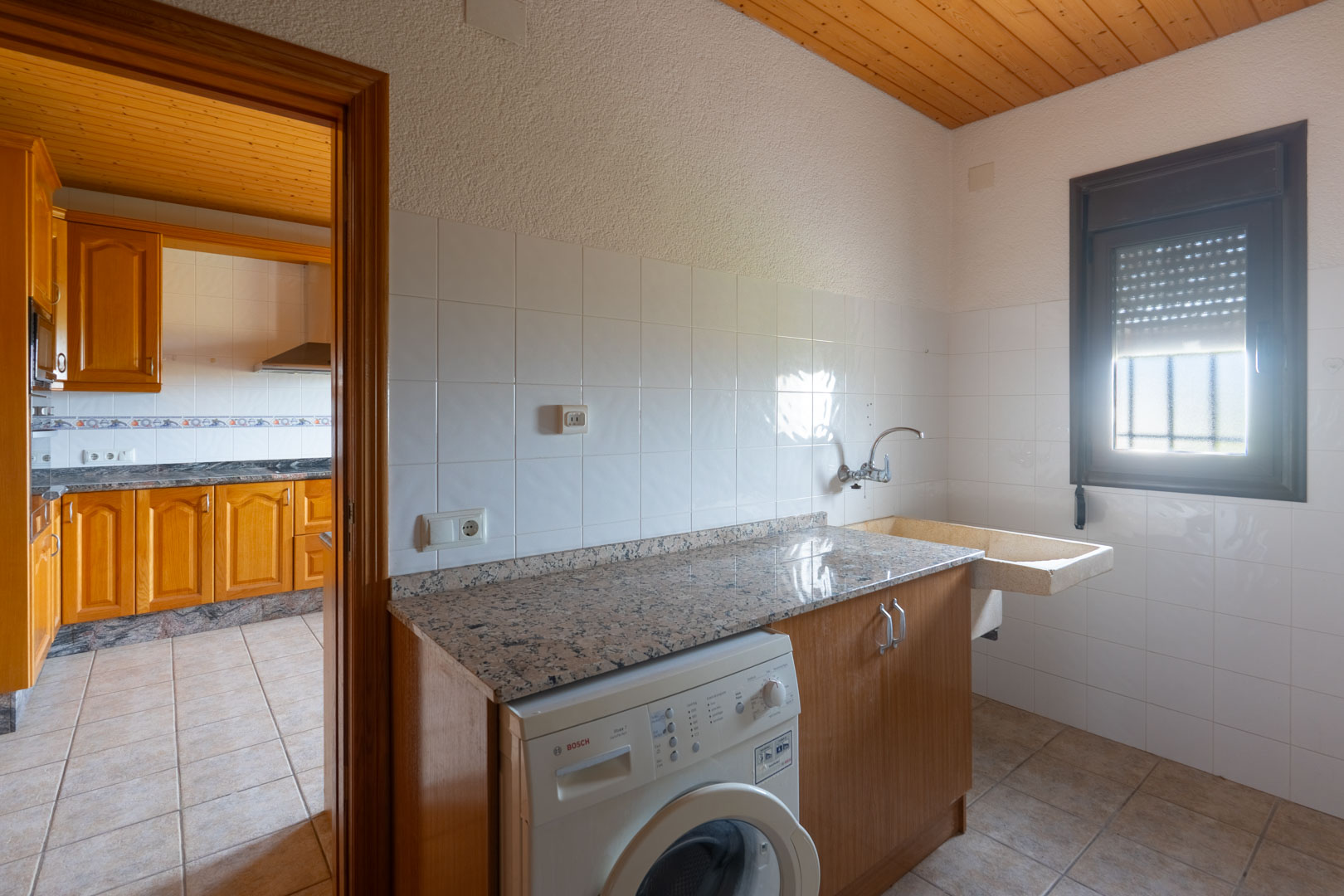 Utility room featuring a granite countertop, Bosch washing machine, and traditional stone sink. The space includes white wall tiles and a wooden ceiling, adjacent to the kitchen.